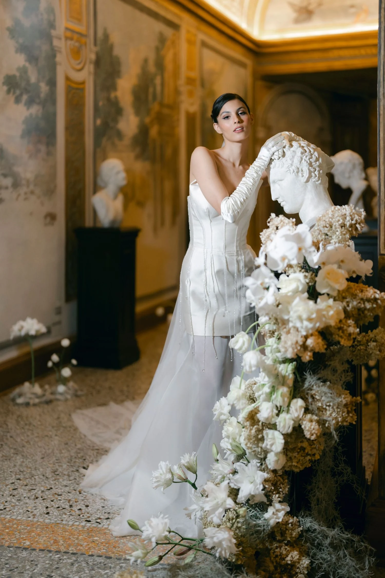 Bride beside a cascading white and yellow floral installation in a frescoed Roman salon