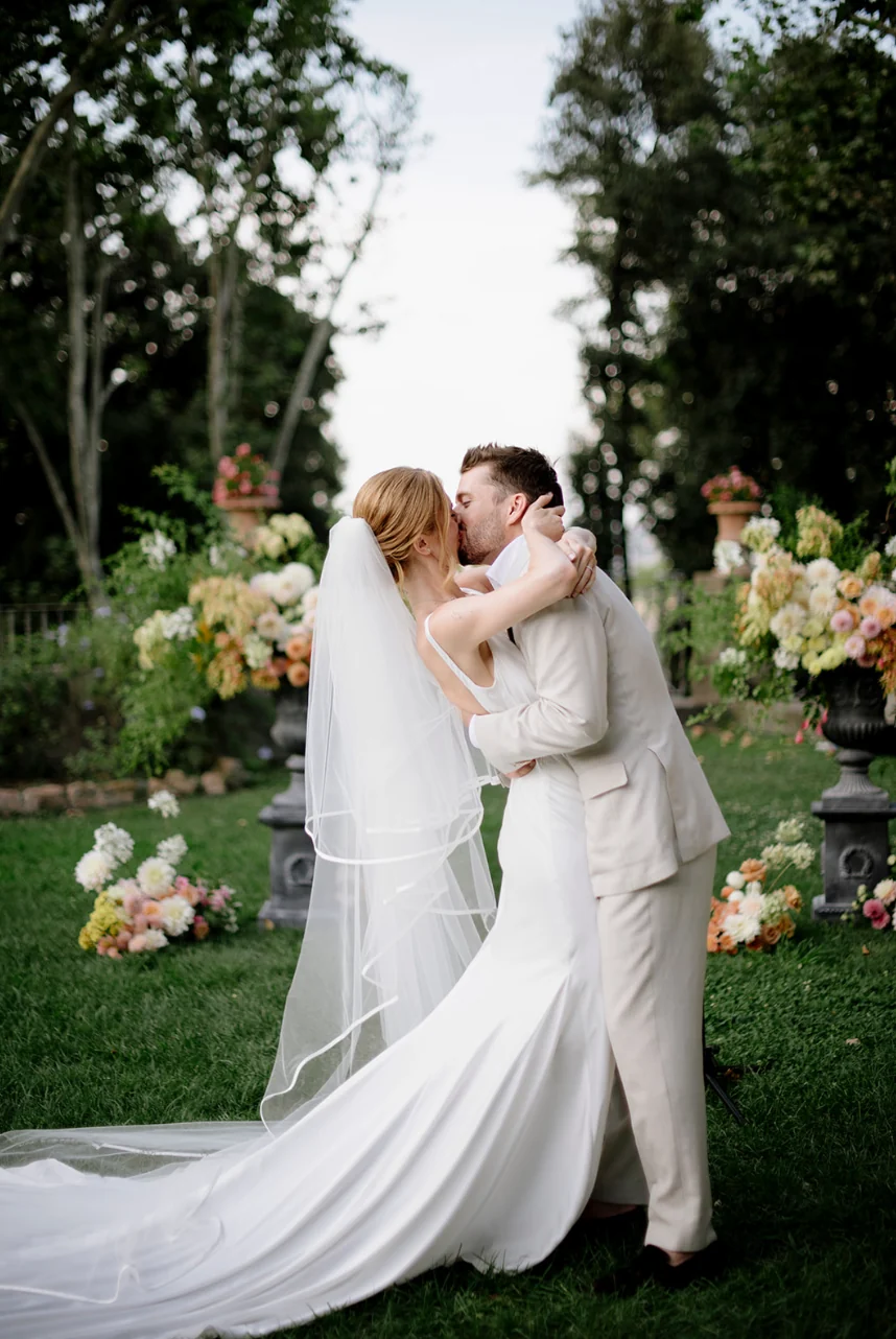 Bride and groom embracing on a lawn between soft peach and cream ground arrangements